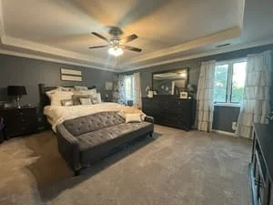Clean master bedroom with made bed, dark furniture, carpet, and windows